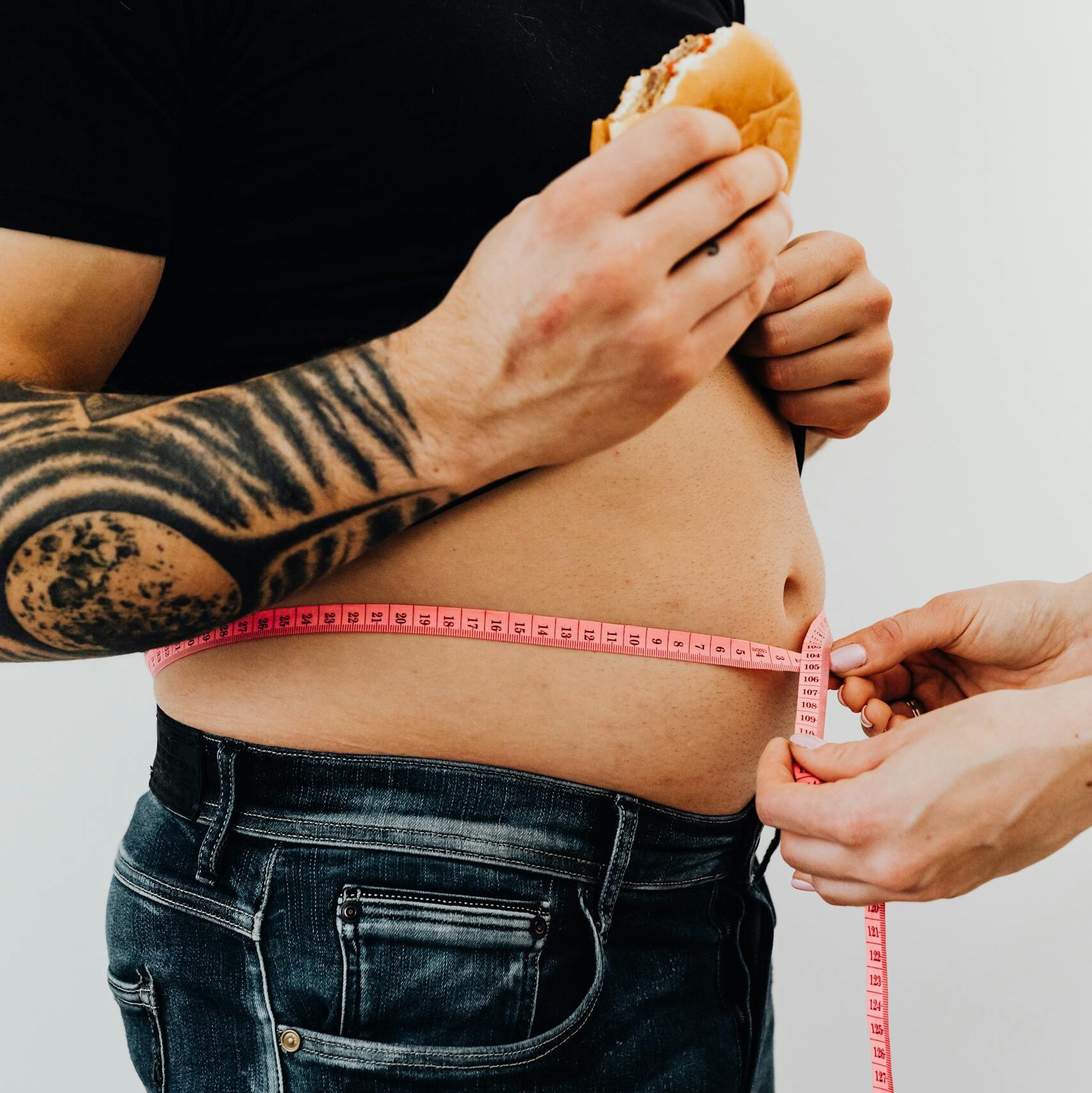 Close-up of a man holding a burger while measuring his waistline with a tape, highlighting unhealthy eating habits.
