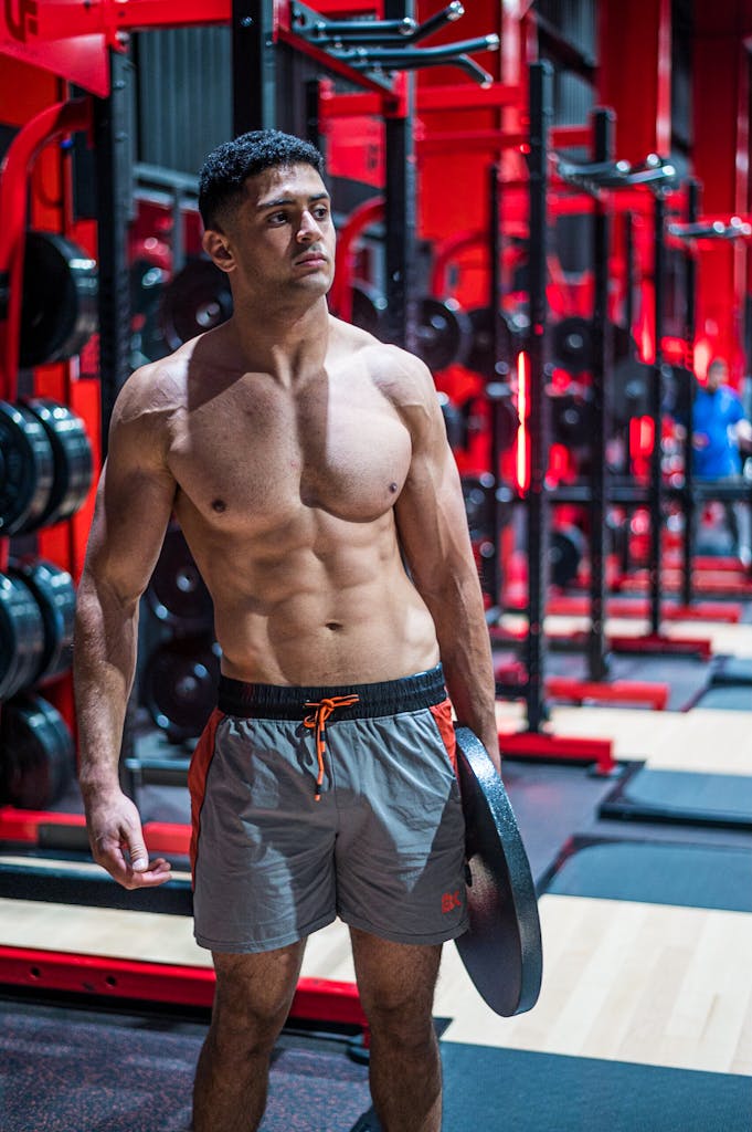 Shirtless man working out with weight plate in a modern gym setting.